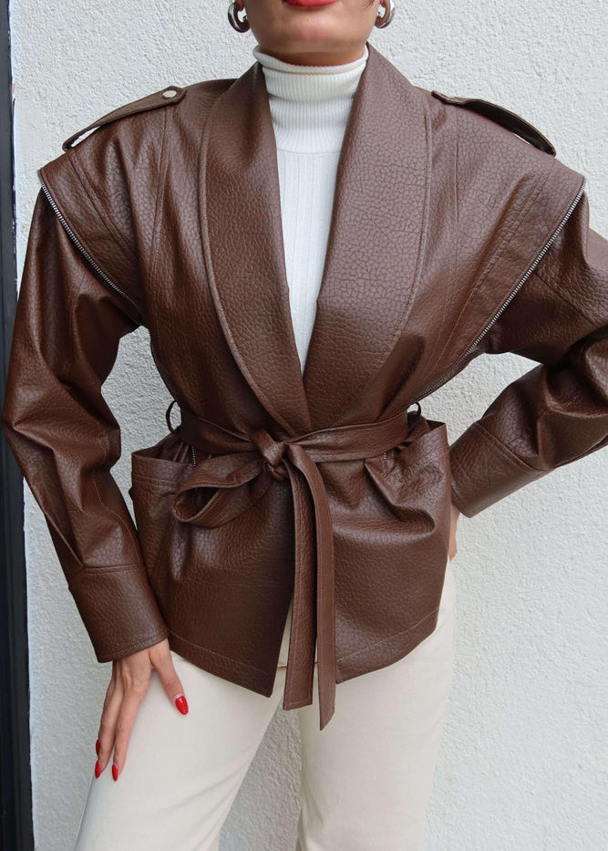 Cette veste femme, d'un riche coloris marron, est confectionnée dans un matériau à l'aspect cuir texturé, lui conférant un style audacieux et sophistiqué. Elle se distingue par sa coupe ceinturée à la taille, qui permet de structurer la silhouette et de créer un effet blouson élégant.
La veste présente un col châle ample et drapé, ajoutant une touche de raffinement. Des détails de pattes boutonnées sur les épaules rappellent le style des vestes motard ou des trenchs, tandis que les zips décoratifs sur le ha